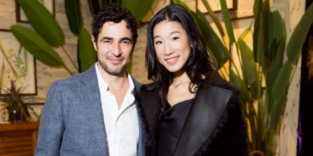 A smiling man in a gray blazer stands next to a smiling woman in a black dress and coat, in front of large green plants that evoke an exotic Paradise. They pose together indoors at an event during the 2025 SF Fall Show. C MAGAZINE is displayed above them in large black letters.
